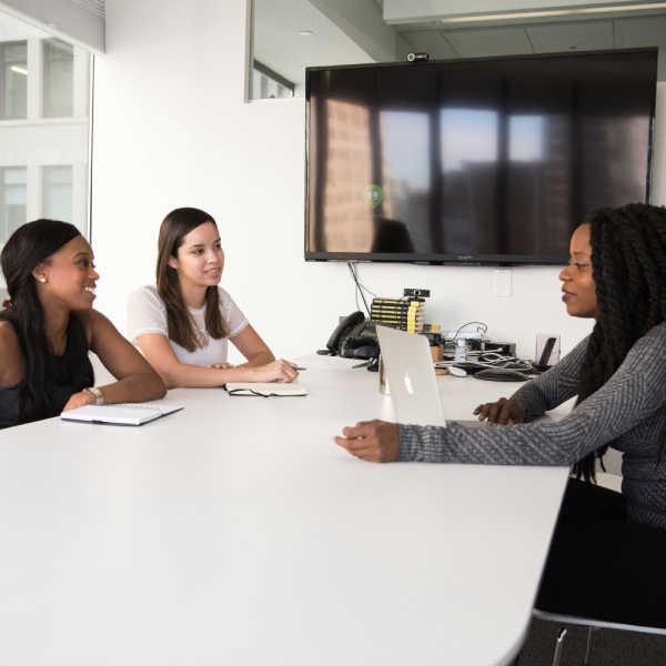 Three women engaged in discussion while seated at a table in a modern office setting.